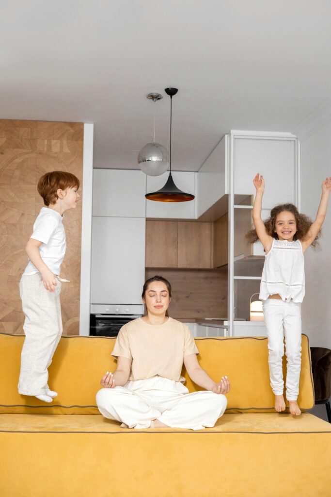 A mother sits meditating on the couch while her children jump around her, illustrating the mental load of motherhood as she seeks moments of calm amidst the chaos of parenting.