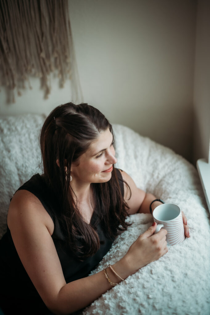 A woman relaxes in a cozy corner, holding a mug and gazing thoughtfully out the window.