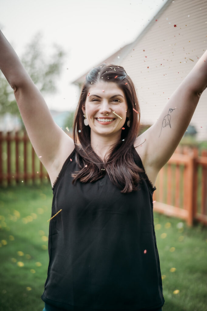 A joyful woman stands outside, arms raised in celebration, surrounded by confetti.