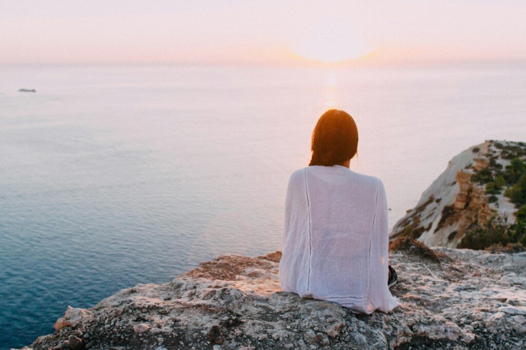 A woman sits on a rocky cliff, gazing at a serene ocean sunset. | EMDR and ERP