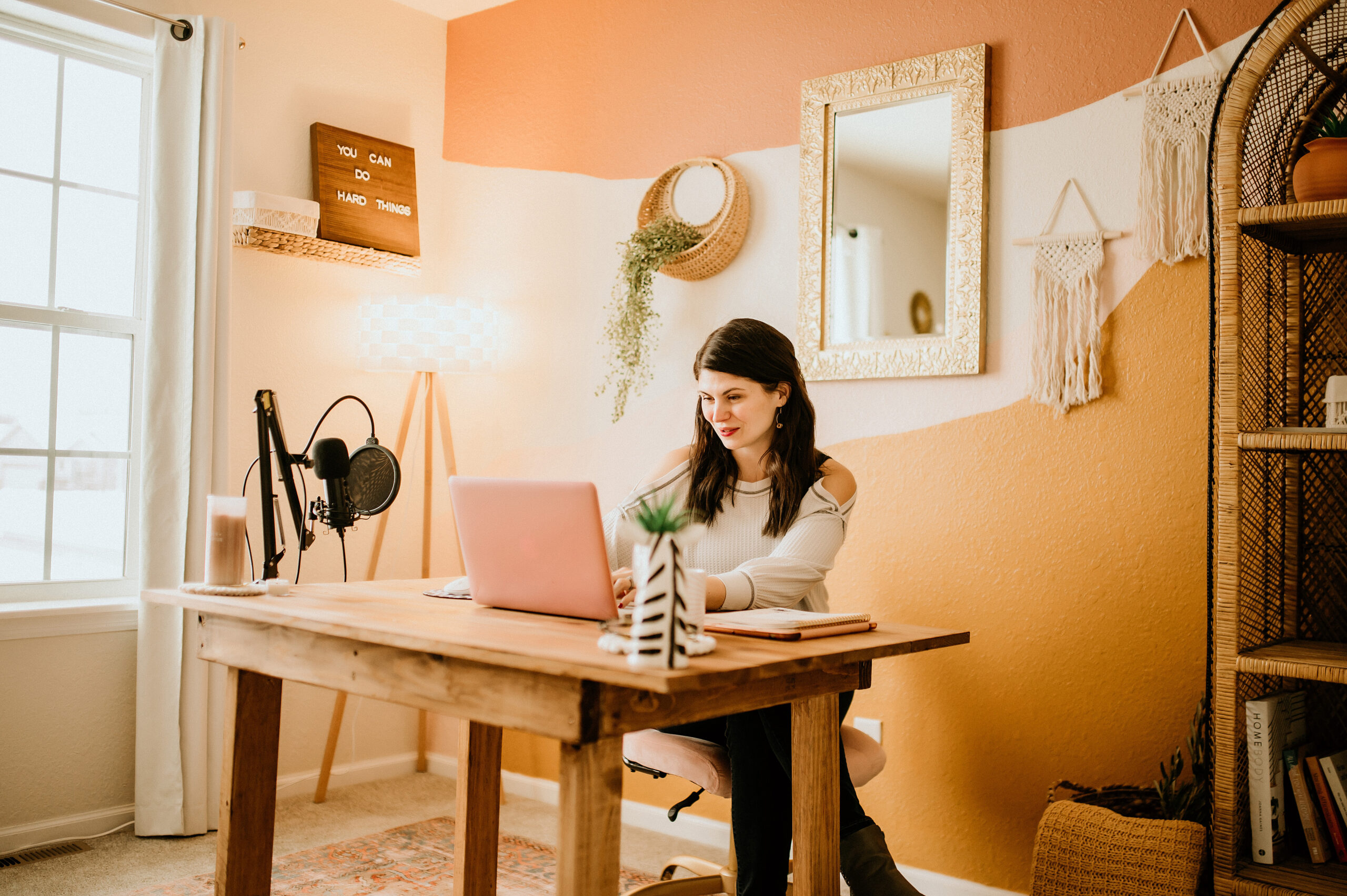 A woman works at a wooden desk with a laptop in a cozy, sunlit office decorated with warm tones, greenery, and motivational wall art.