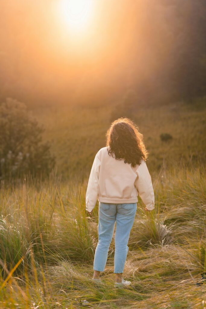 A young girl stands in a grassy field, facing a golden sunset. Wearing light jeans and a cream sweater. The serene scene captures a peaceful connection with nature even when managing OCD in kids.