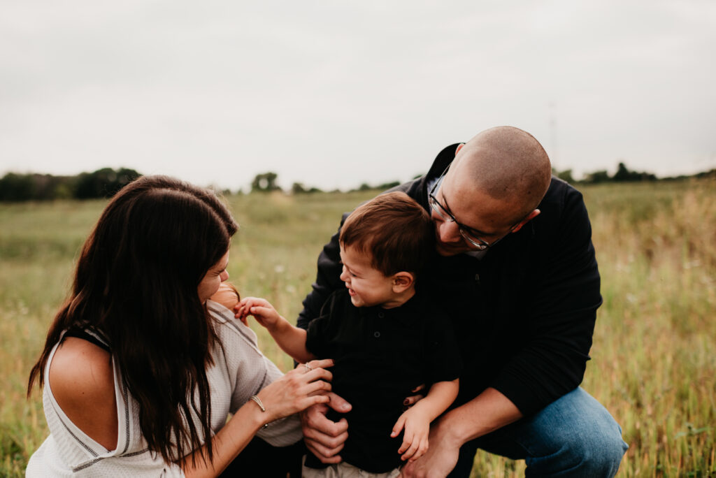 A joyful family moment in a grassy field. A laughing child in a black shirt is lovingly embraced by his parents, who lean in with warm smiles. 