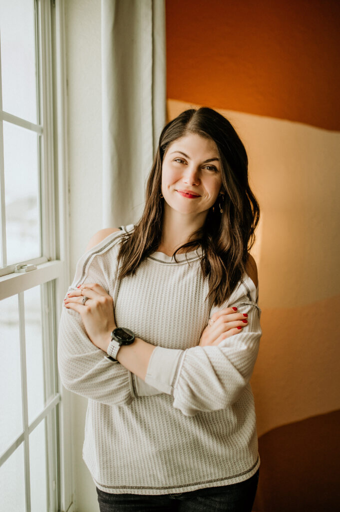 A confident woman smiles warmly while standing by a window in a cozy room. Her relaxed demeanor exudes positivity, embodying resilience and the strength to face challenges head-on.