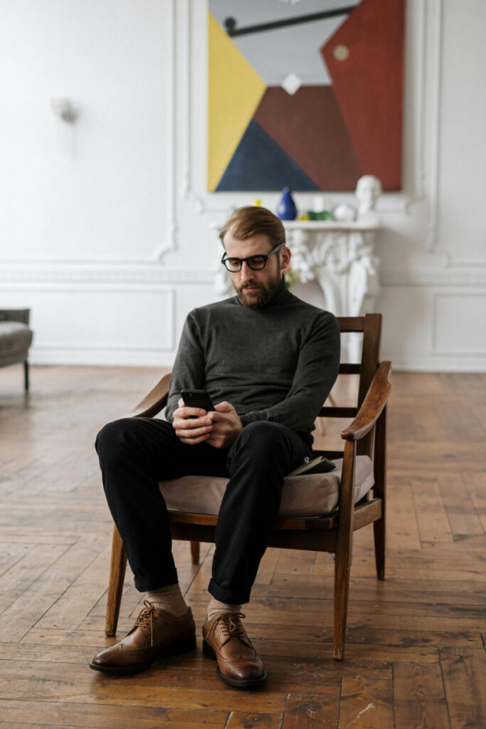 A man sits in a modern room looking at his phone, illustrating the connection between screen time and how technology affects mental health.