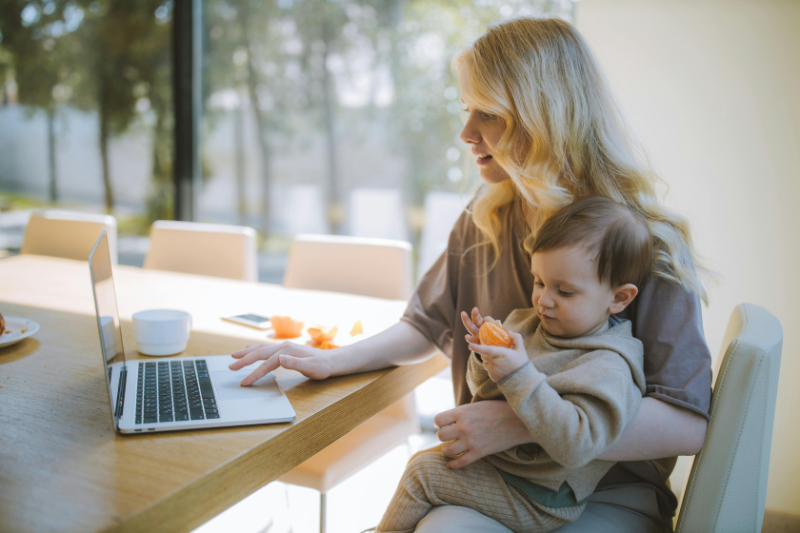 A working parent balances managing anxiety and OCD while working on a laptop with a toddler in their lap, multitasking in a bright kitchen.