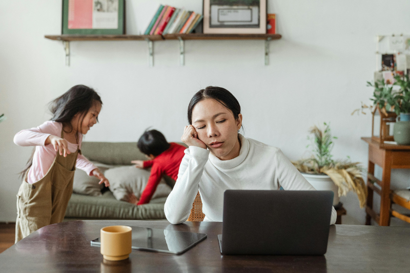 A tired parent sits at a laptop while children play energetically in the background, illustrating the stress of managing anxiety and OCD as a working parent.