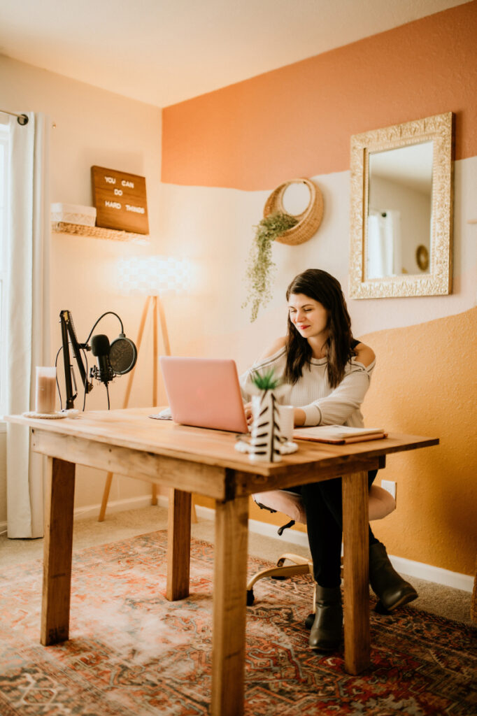 A woman smiles while working on a laptop in a cozy home office with a sign reading "You can do hard things" in the background.