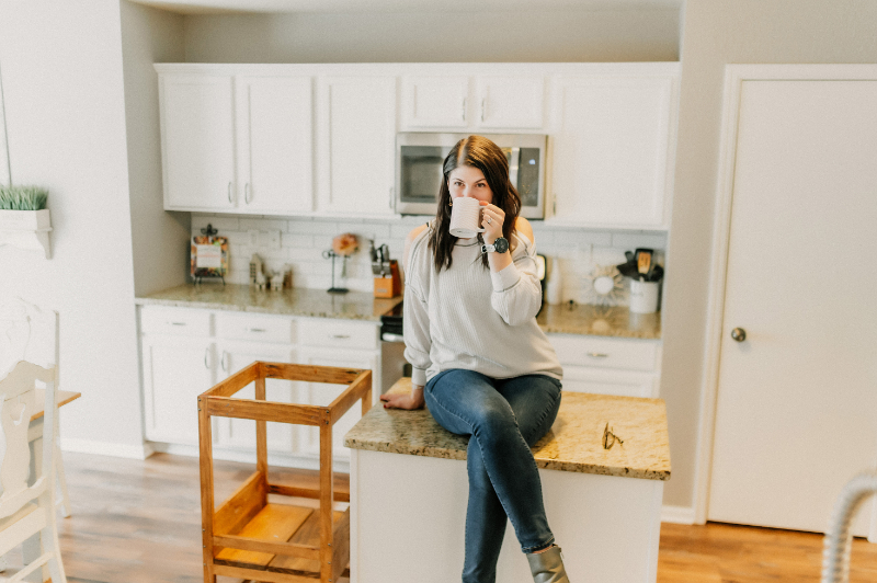 A woman sits on a kitchen counter, sipping from a mug, in a bright, white kitchen with modern decor.