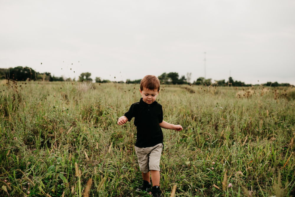 A young boy walks through a tall grassy field on a cloudy day, looking down with curiosity and calm as he explores the open nature around him.