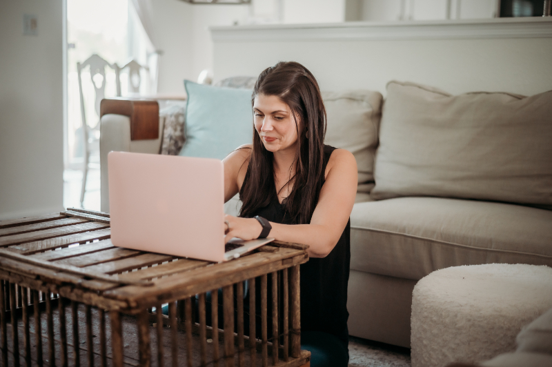 woman sits on the ground in her living room working on pink laptop