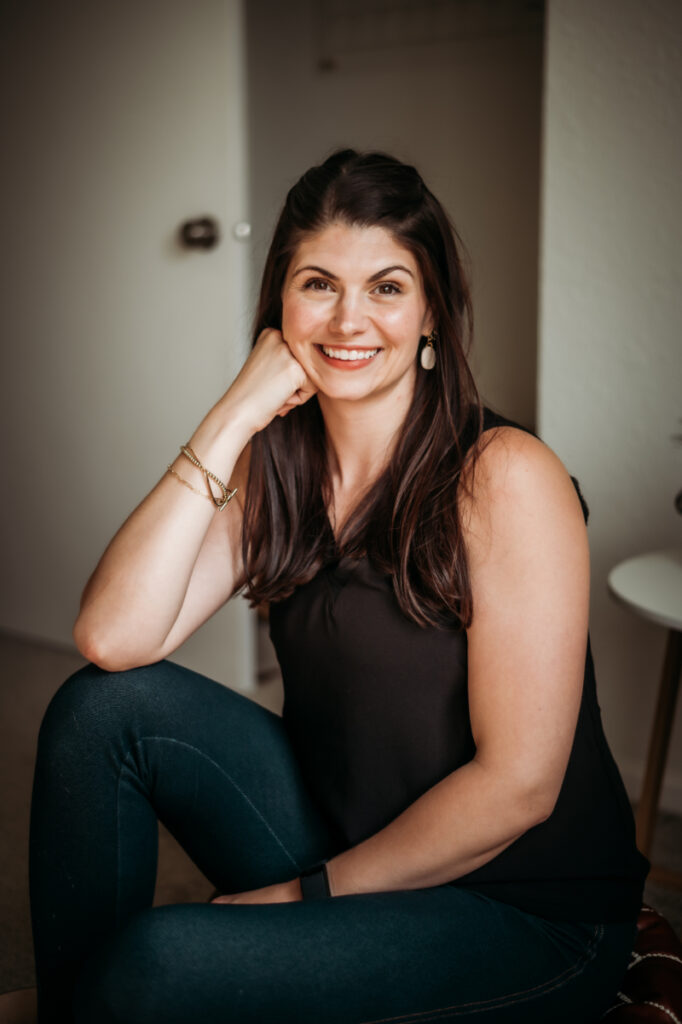 Woman sits casually on the floor, smiling warmly with a relaxed, friendly expression.