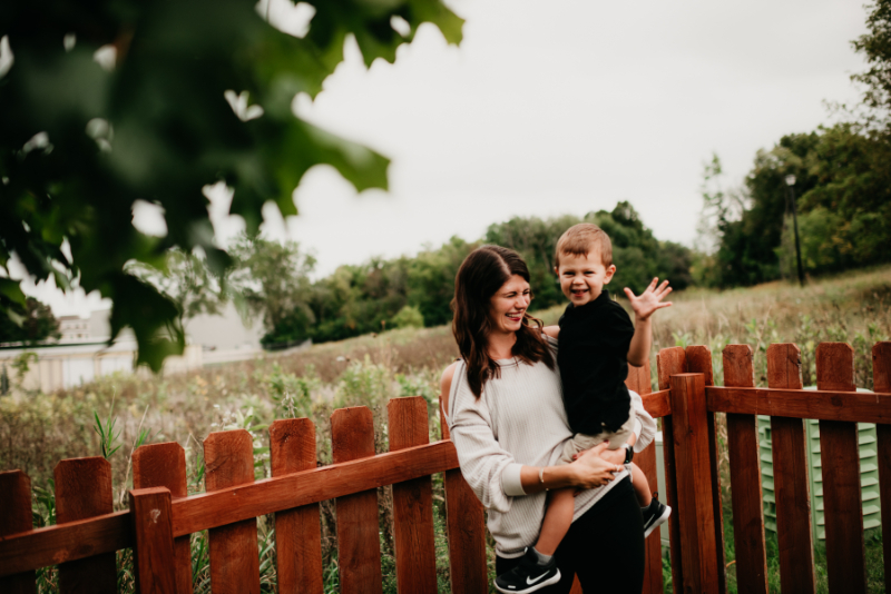 Woman holds smiling child by a wooden fence in a peaceful outdoor setting.