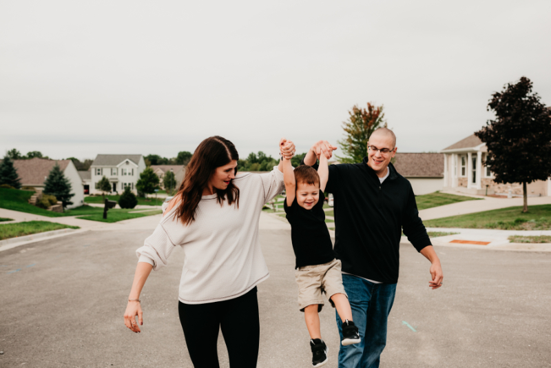 Family walks on a suburban street, parents swinging child playfully between them.