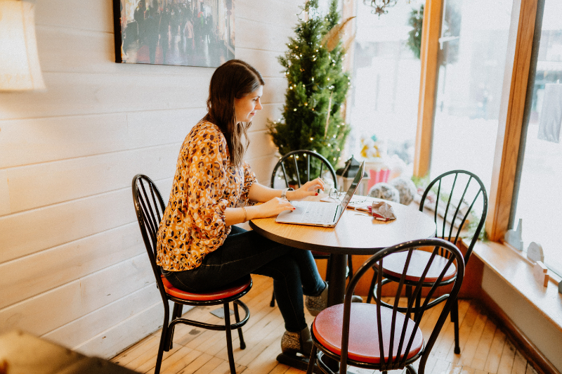 Woman works on a laptop at a cozy cafe table, decorated with holiday greenery by the window.