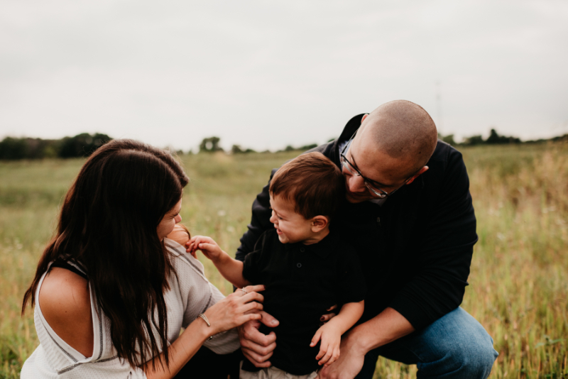 Family kneels in a field, parents smiling and engaging with their young child.