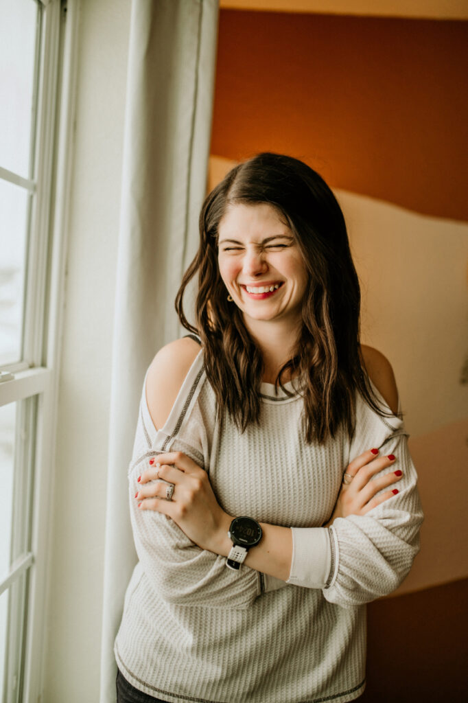 Woman laughs joyfully while standing by a window, arms crossed, wearing a light sweater.