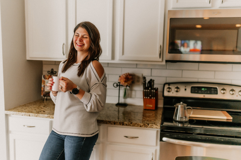 Woman smiles while holding a cup of coffee, standing in a bright kitchen with white cabinets.