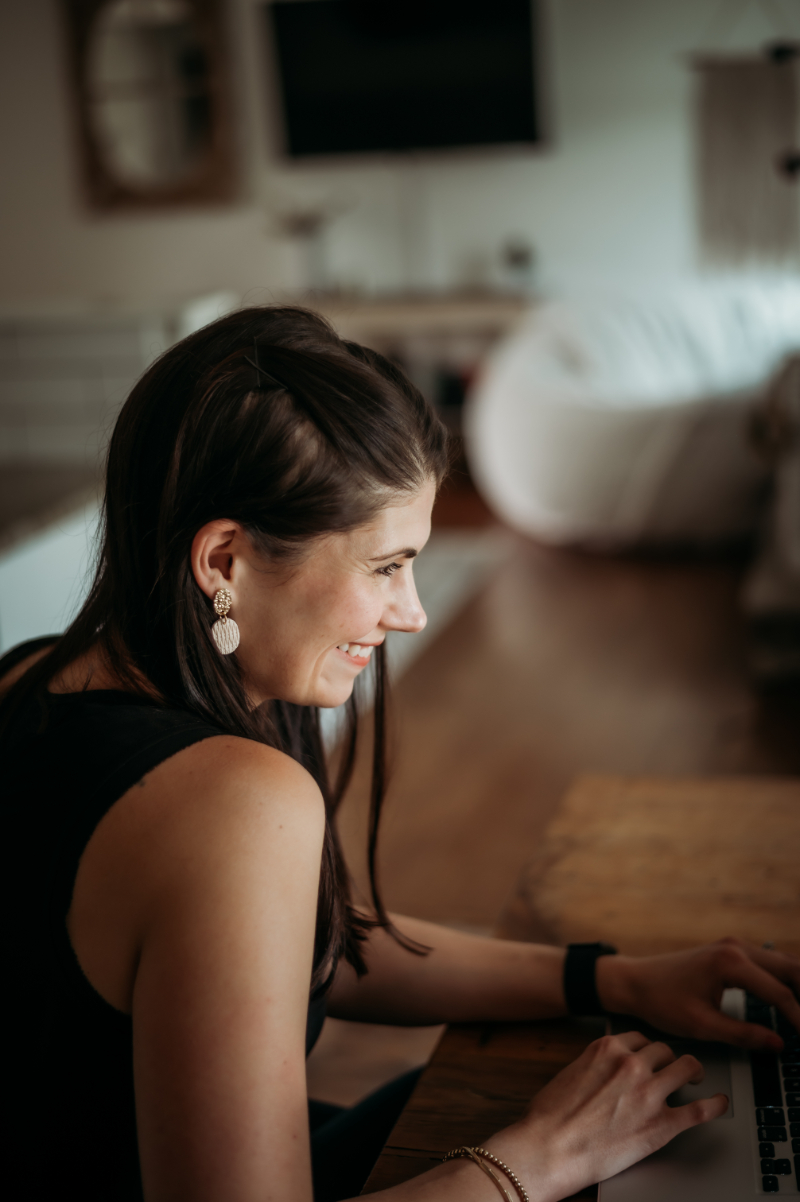 Close-up of woman smiling while typing on a laptop at a wooden table in a cozy living room.