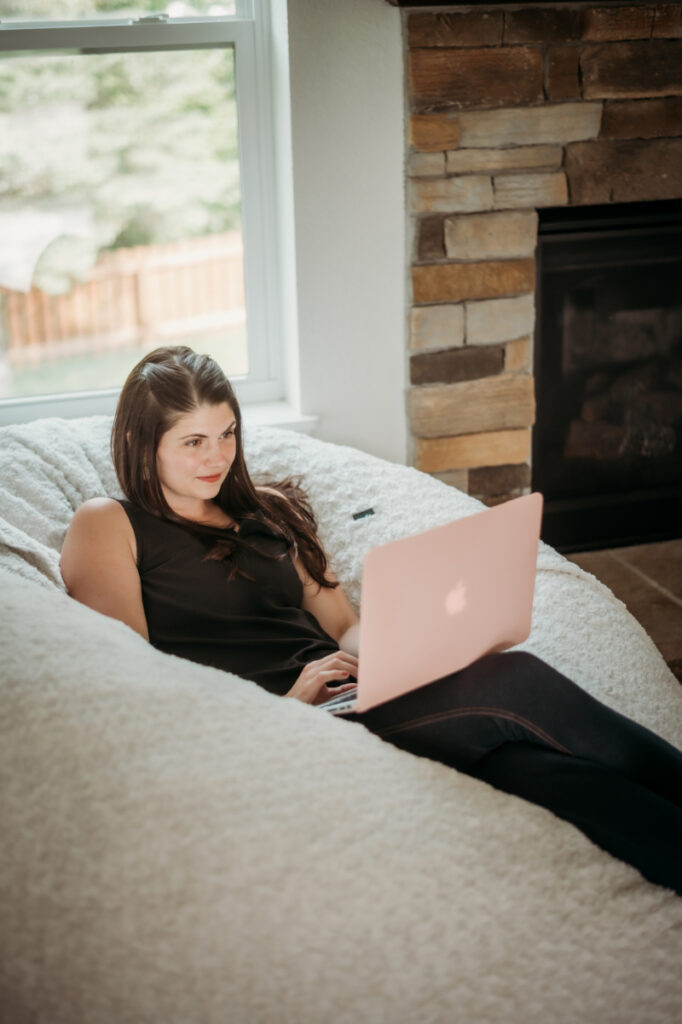 Woman relaxes on a cozy bean bag chair, working on a pink laptop near a stone fireplace.