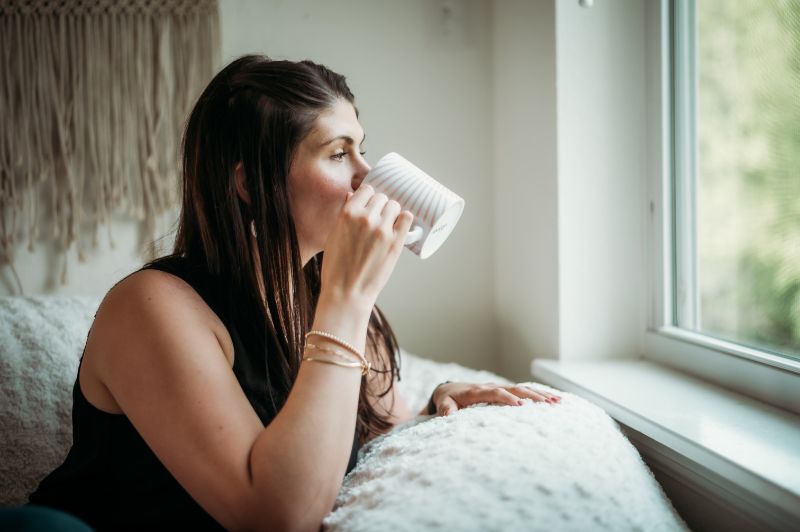 A woman sips coffee by the window, deep in thought—reflecting on parenting and mental health in a quiet moment.