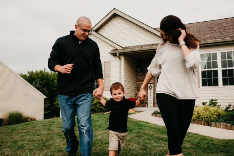 Parents hold hands with their child, walking outside their home—joyful parenting and mental health in motion.