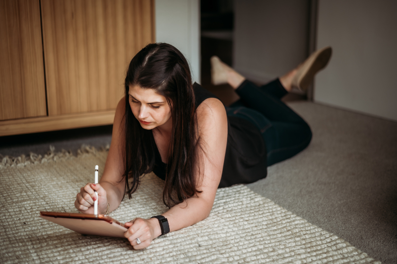 A woman lies on the floor using a digital tablet, possibly planning or journaling—taking time for reflection during summer break while managing OCD routines and goals.