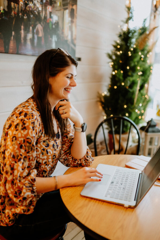 A smiling woman works at a laptop in a cozy, festive café, appearing focused and engaged.