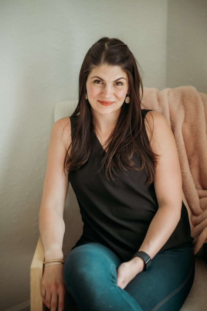A woman sits relaxed in a soft chair, gently smiling—offering a calm, supportive presence.