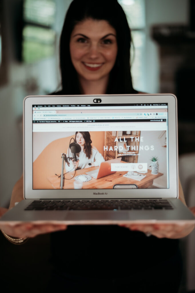 A smiling woman holds a laptop forward, displaying her podcast “All The Hard Things,” which offers real talk on topics like ADHD and OCD recovery and parenting.